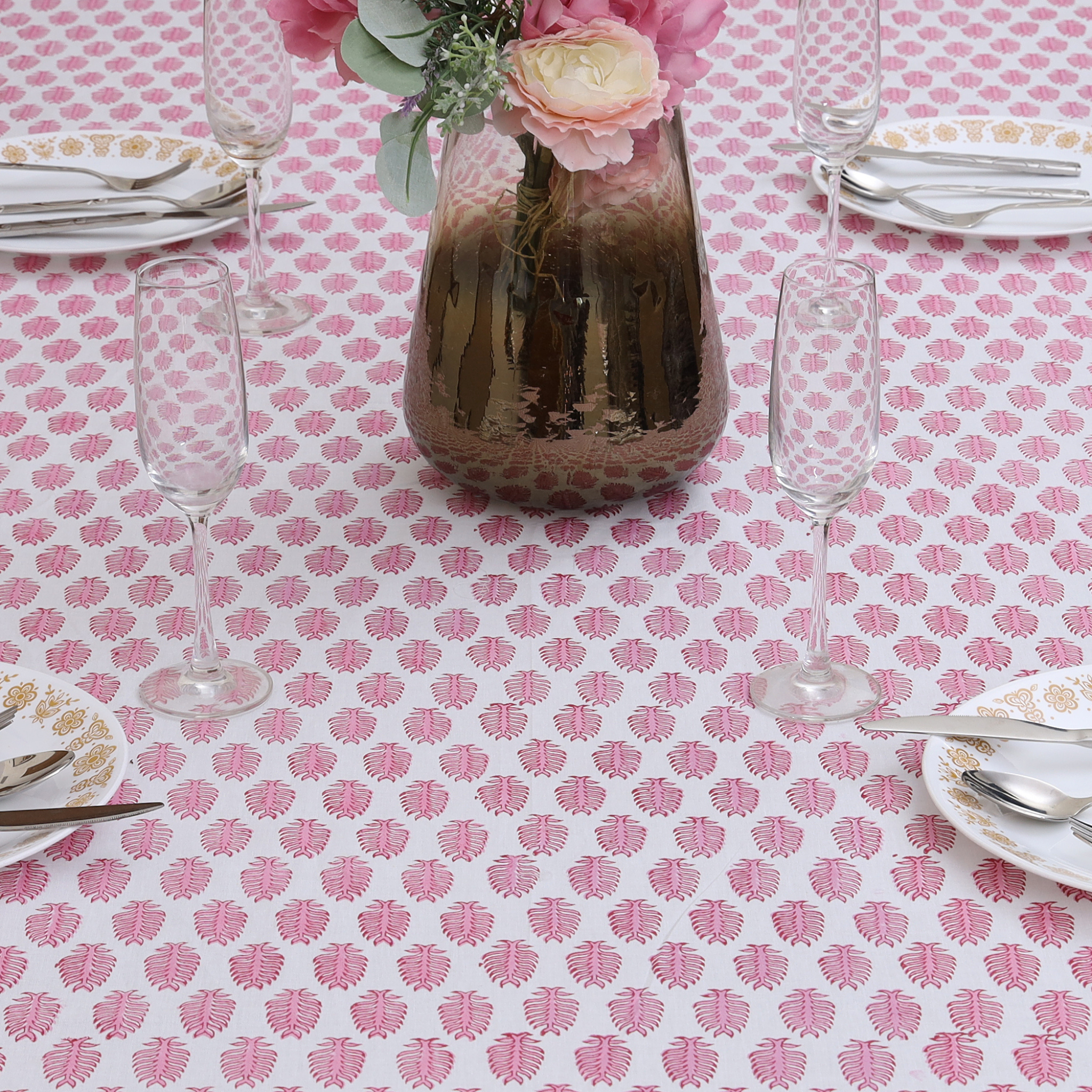 Table setting with a pink floral patterned tablecloth, glasses, plates, and a vase with flowers.