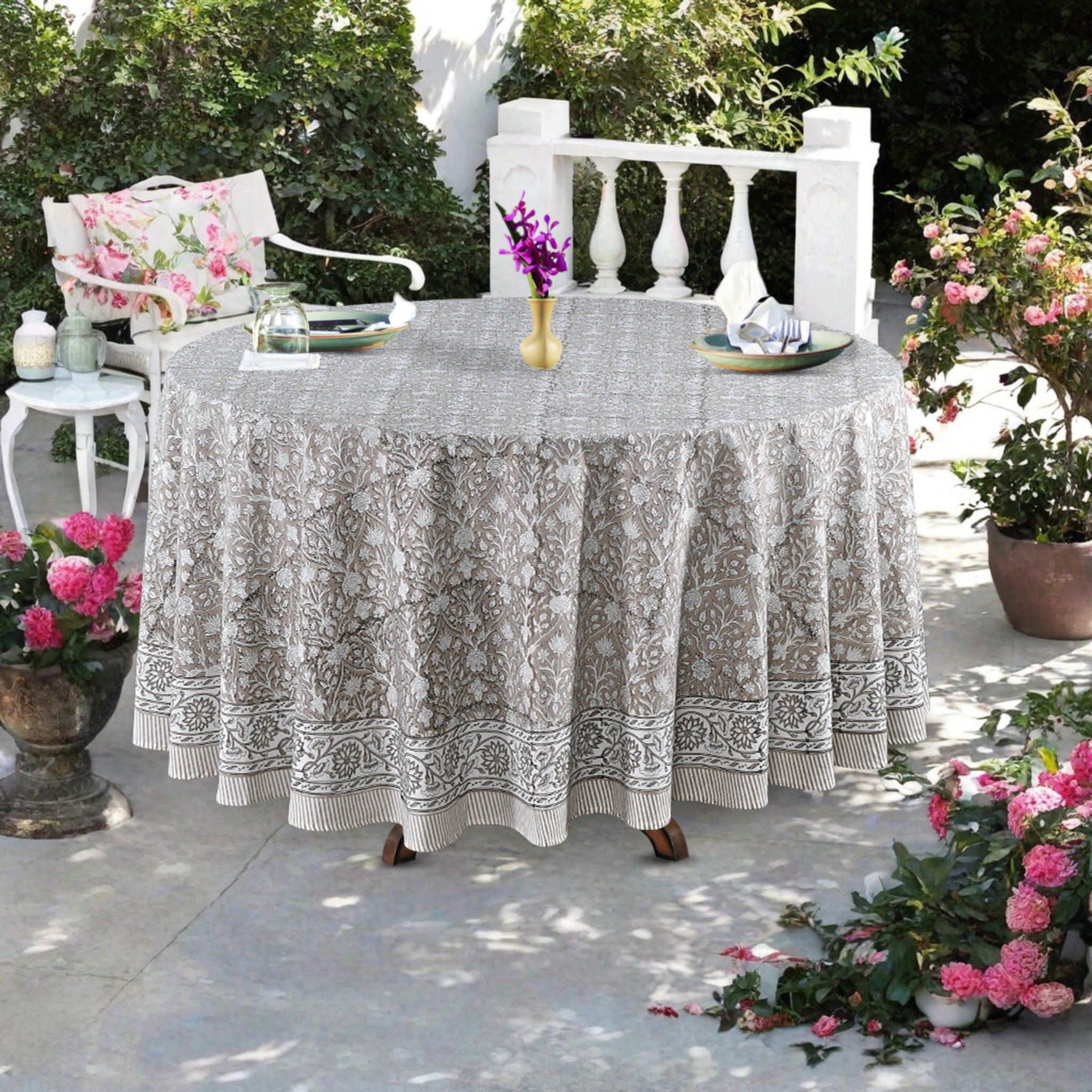Round table with a patterned tablecloth outdoors, surrounded by potted plants and chairs.