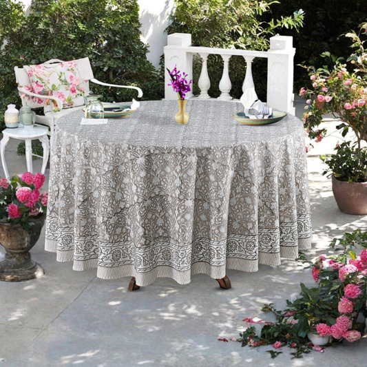 Round table with a patterned tablecloth outdoors, surrounded by potted plants and chairs.
