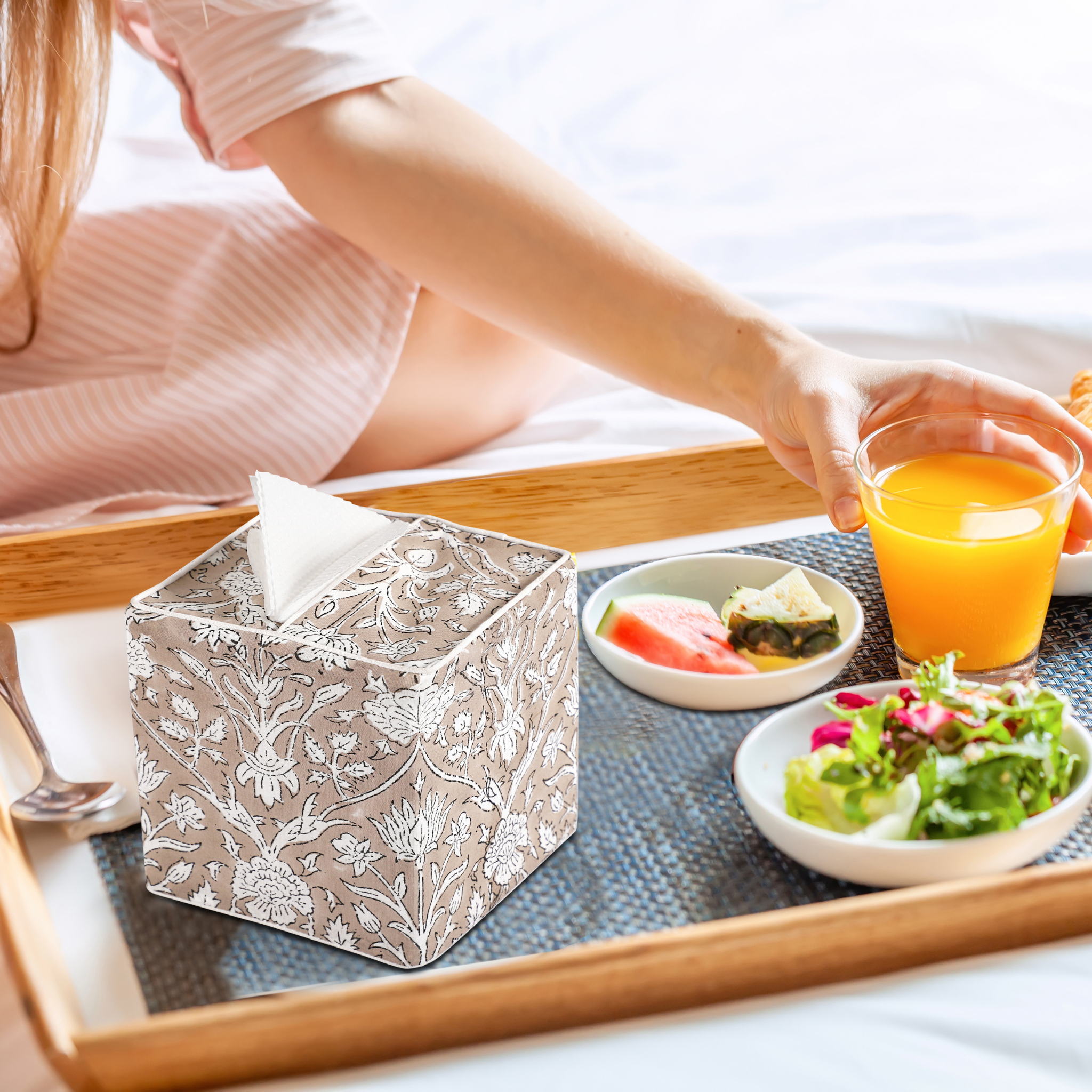 Person holding a glass of orange juice with a floral-patterned tissue box on a tray.