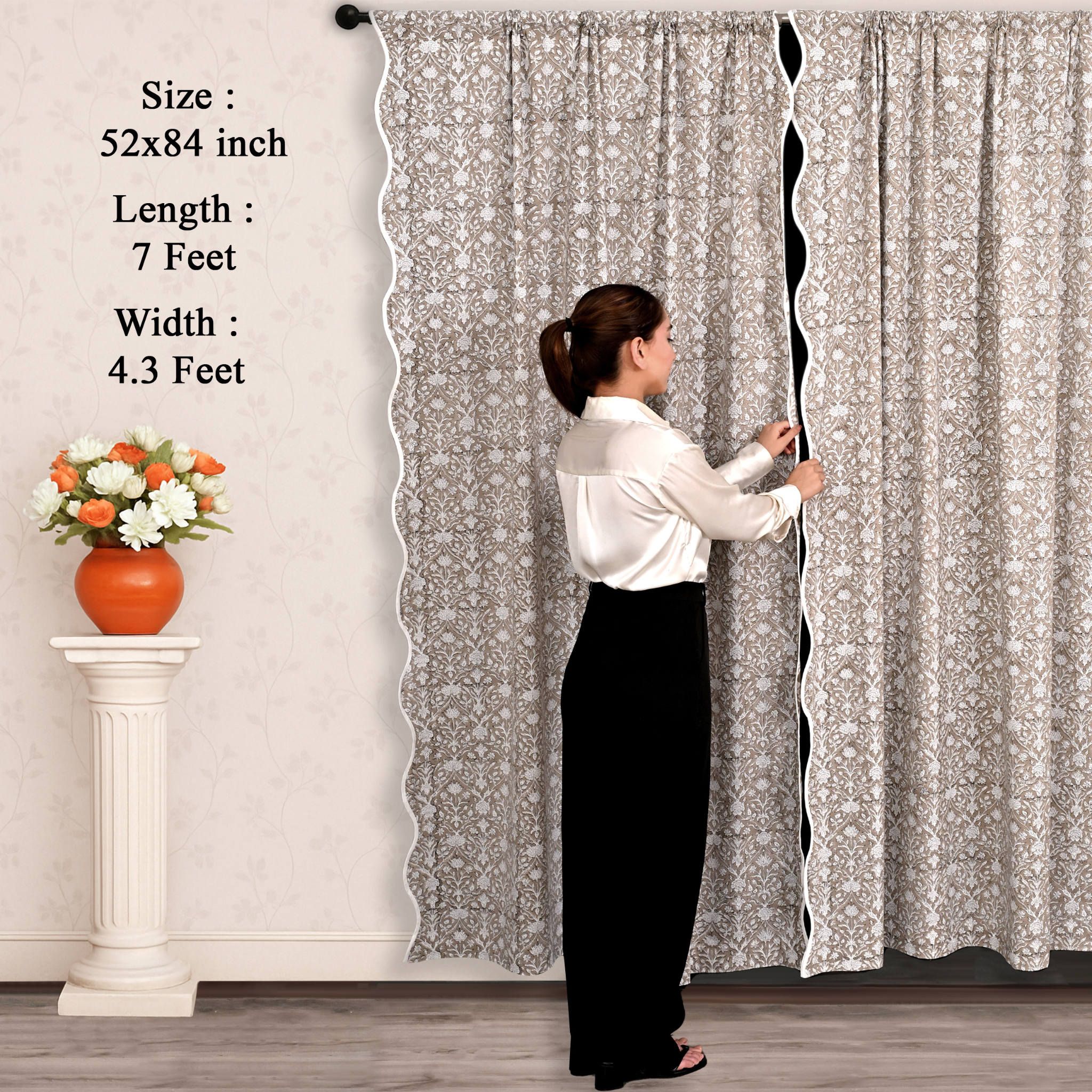 Person adjusting a long curtain with floral pattern, next to a small table with flowers.