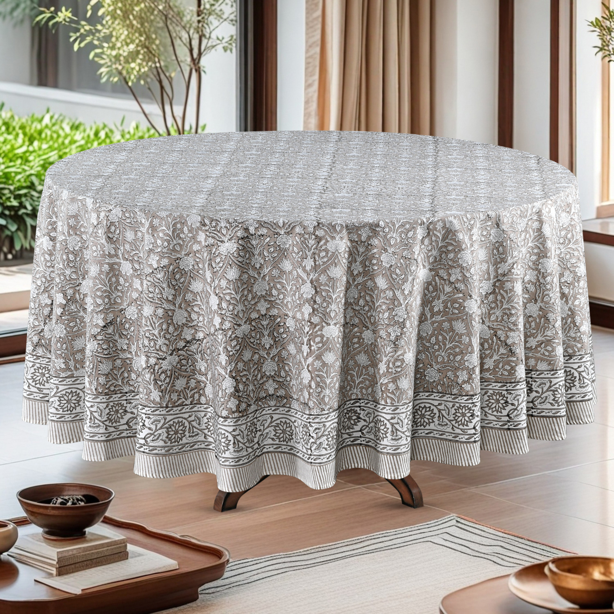 Round table with a patterned tablecloth in a bright room with plants and curtains.