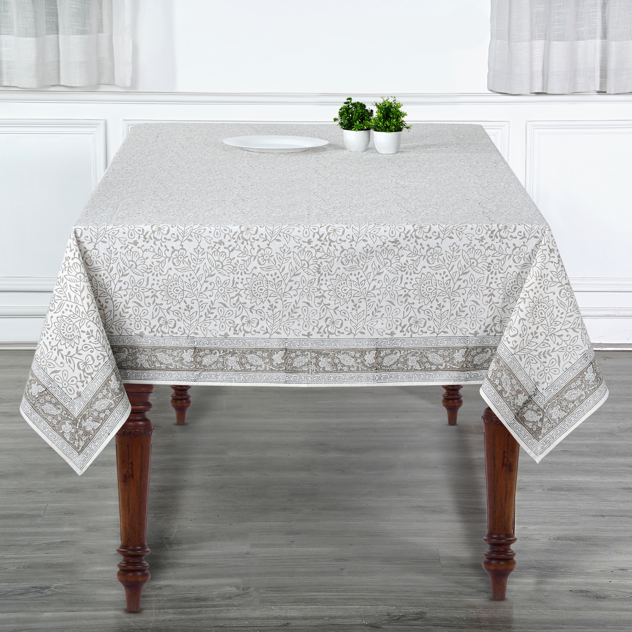 Tablecloth with floral pattern on a wooden table in a room with white walls and gray floor.