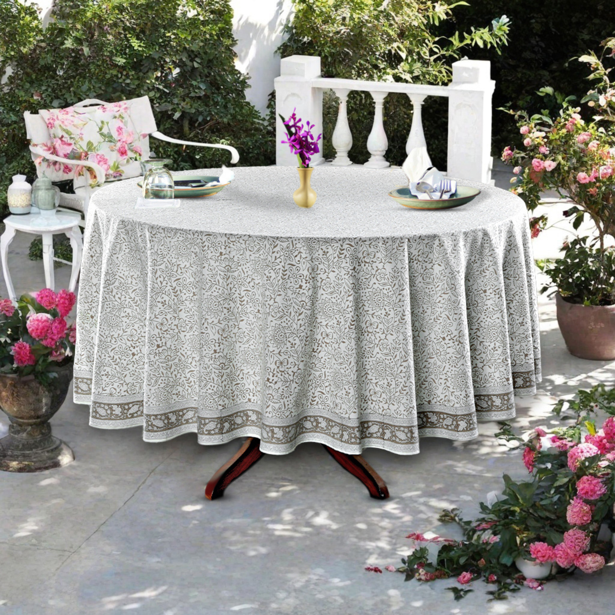 Round table with a decorative tablecloth in an outdoor setting with flowers and chairs.