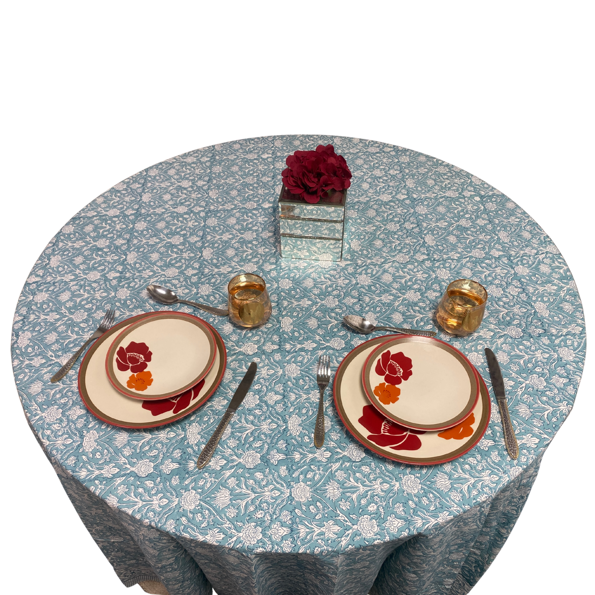 Round table with floral tablecloth, plates, cutlery, and centerpieces on a white background