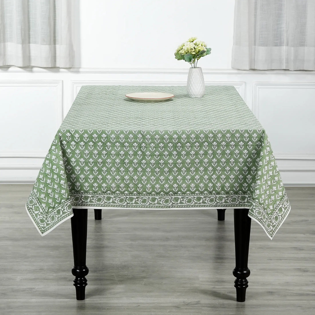 Green patterned tablecloth on a wooden table with a vase of flowers in a room.