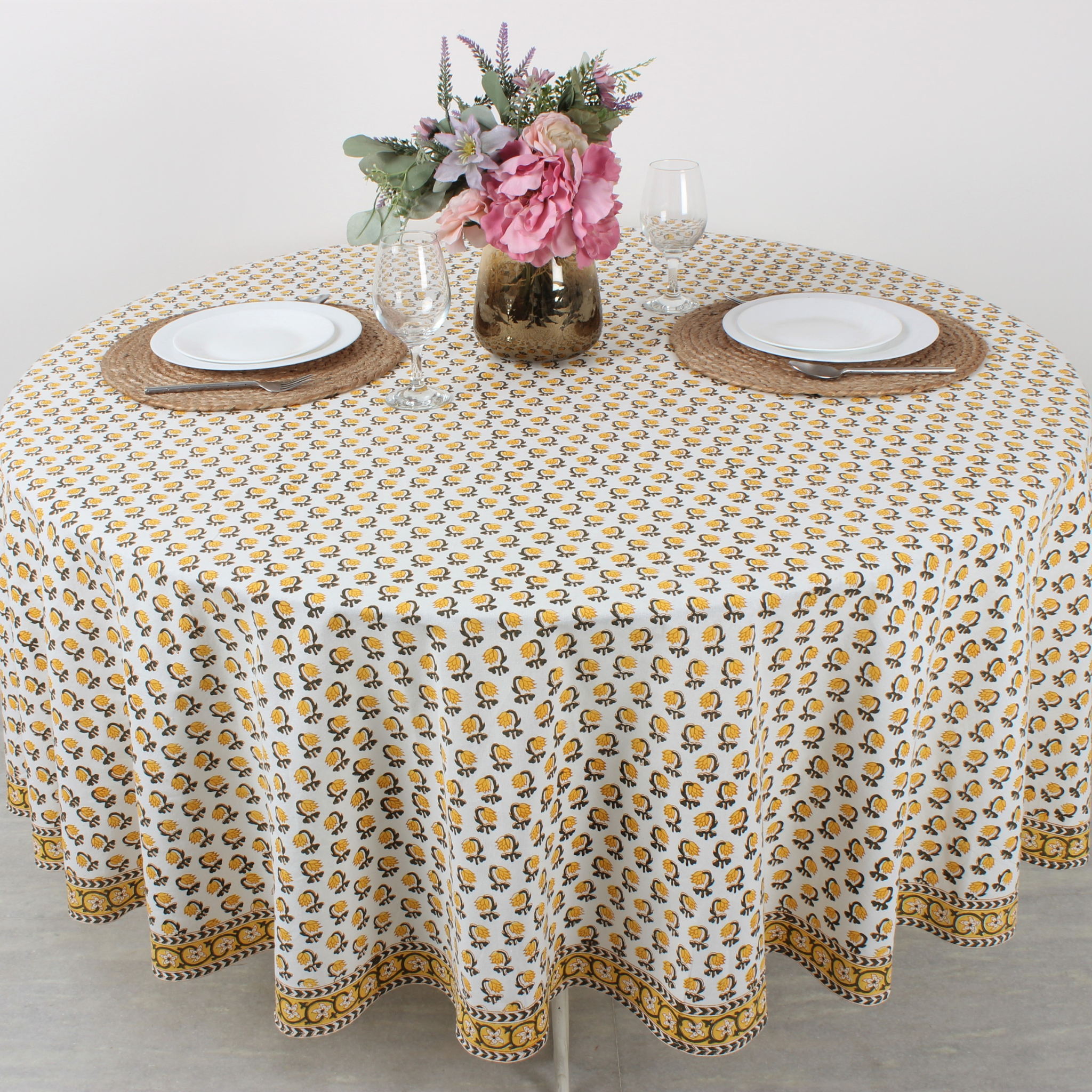 Round table with a floral patterned tablecloth, plates, and flowers on a light background