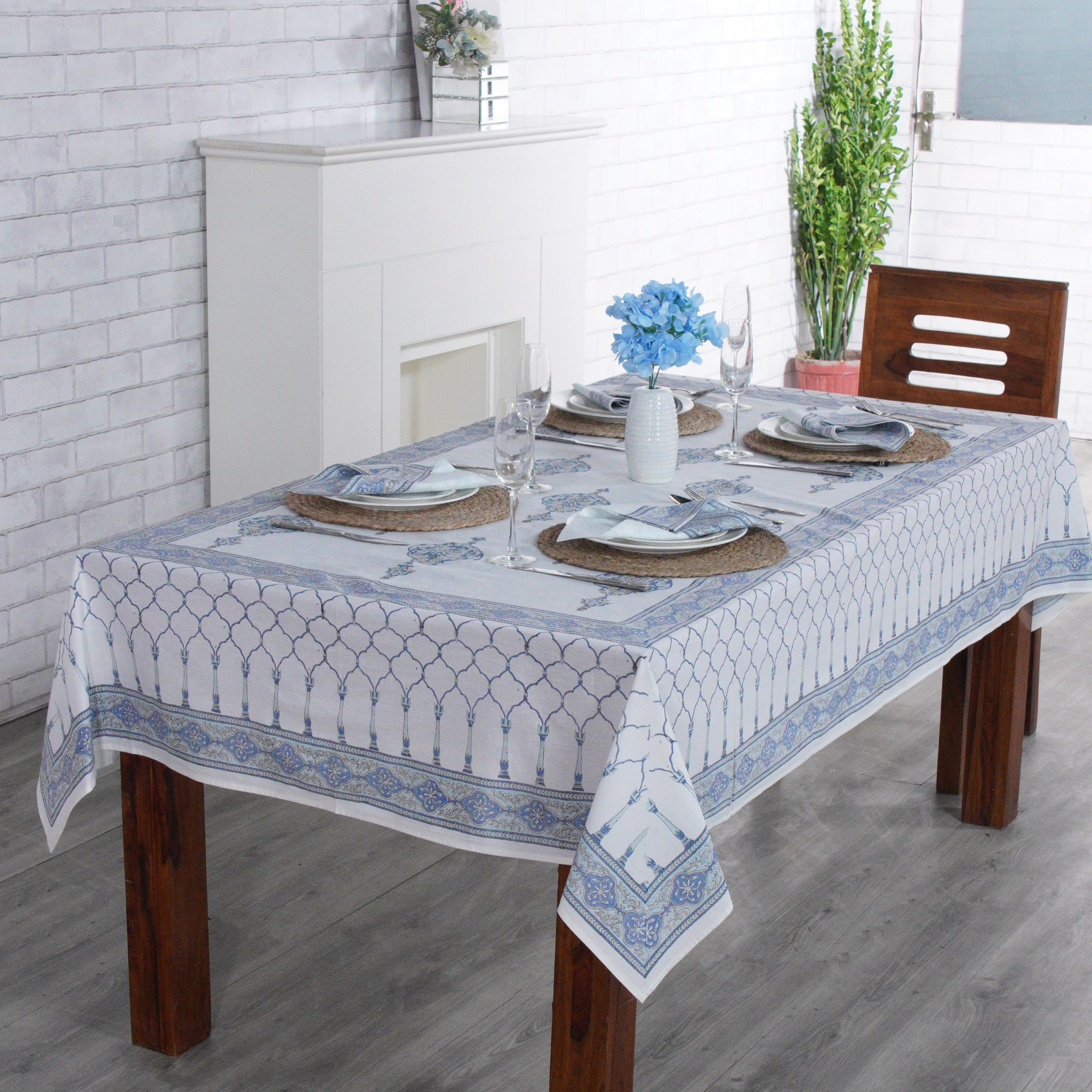 Dining table set with a patterned tablecloth in a room with white walls and wooden floor.
