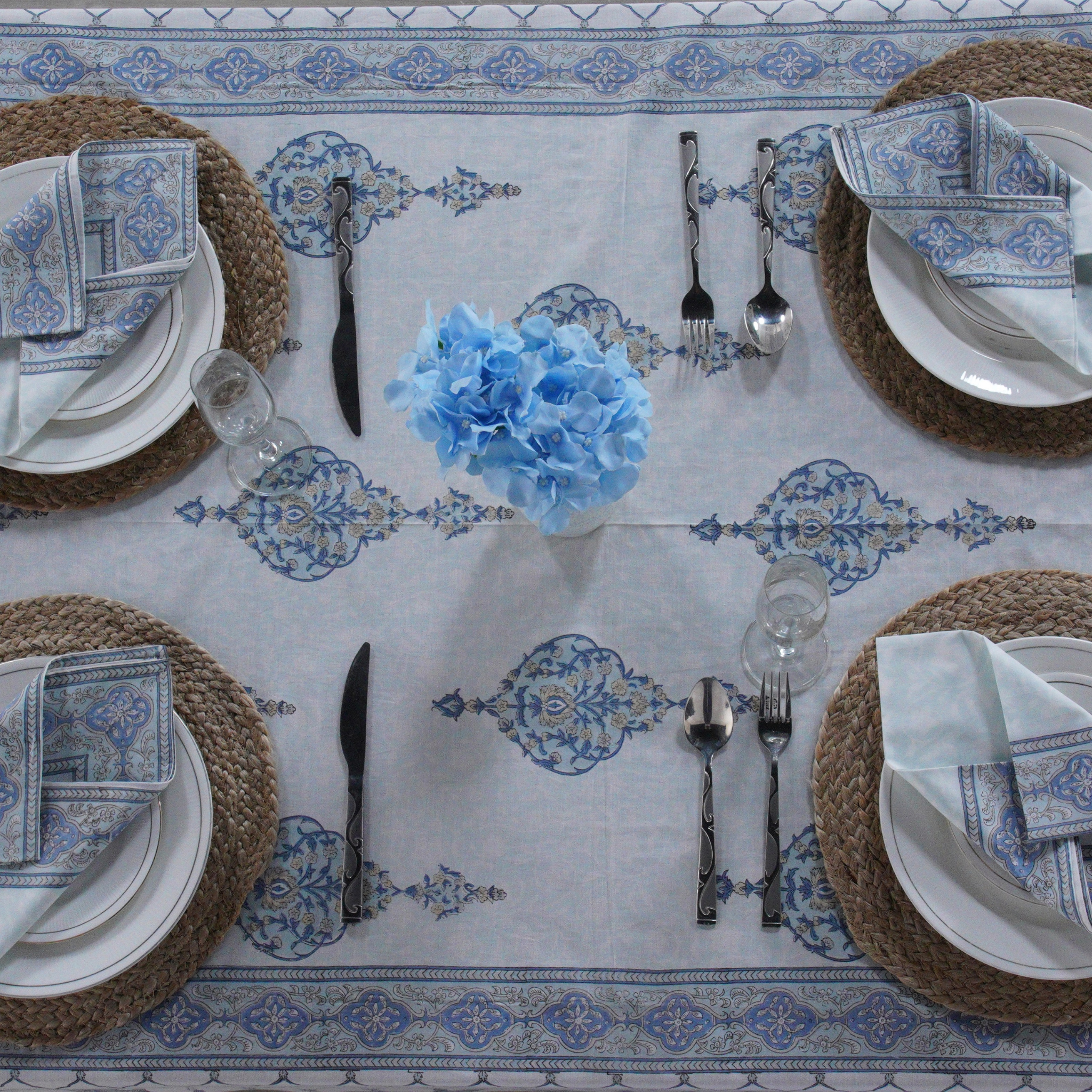 Elegant table setting with blue and white patterned tablecloth, blue flowers, and wicker chargers.